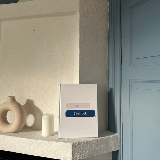 Vit chattbok med texten ’Vår Chattbok’ placerad på en hylla bredvid en vas och ett ljus - en personlig minnesbok av dina konversationer.